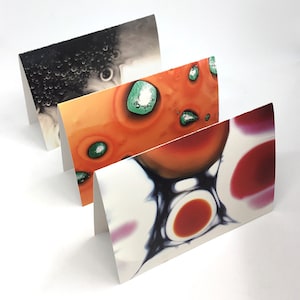 May include: Three folded greeting cards with abstract designs. The top card features a black and white image of bubbles. The middle card has an orange and green abstract design. The bottom card has a red, orange, and purple abstract design.