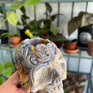 May include: A decorative skull-shaped planter with intricate floral carvings. The planter is a mix of gray, blue, and orange hues, adding a unique touch to any space. Perfect for small plants or succulents.