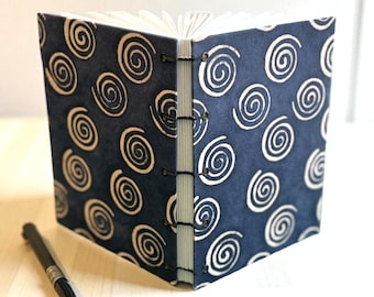 May include: A handmade journal with a navy blue cover featuring a white spiral pattern. The journal is open, revealing blank pages. A black pen rests beside the journal, suggesting its use for writing or sketching.