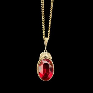 May include: A gold-toned necklace with a large, oval, red gemstone pendant. The pendant is framed with a twisted rope design and adorned with leaf accents. The chain is a delicate, linked style, set against a black background.