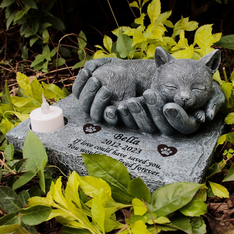 May include: A gray stone memorial with a sleeping cat sculpture. The memorial has a space for a candle and is inscribed with the text "Bella 2012-2023 If love could have saved you, you would have lived forever."