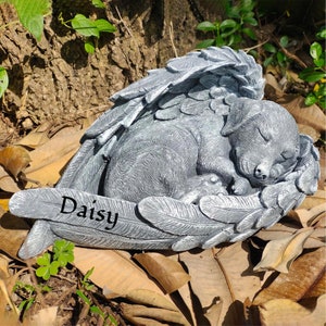 May include: A gray stone memorial statue of a sleeping puppy nestled in angel wings. The name "Daisy" is inscribed on the base. The statue is surrounded by fallen leaves and greenery.