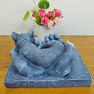 May include: A gray stone sculpture of a cat sleeping in a pair of hands. The sculpture is on a square base with the word "JINHUGBA" printed on the bottom.