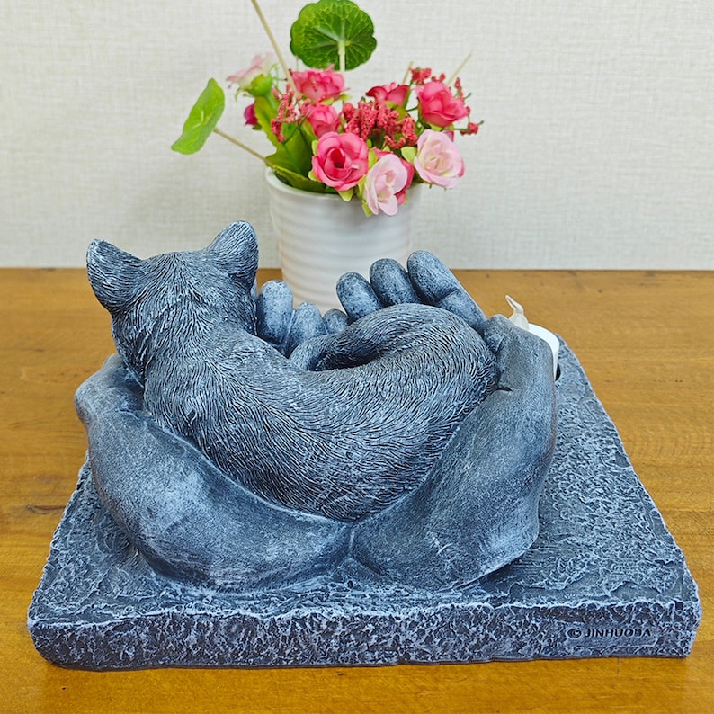 May include: A gray stone sculpture of a cat sleeping in a pair of hands. The sculpture is on a square base with the word "JINHUGBA" printed on the bottom.