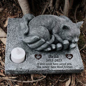 May include: A gray stone memorial with a sleeping cat sculpture in a pair of hands. The memorial has the inscription "Bella 2012-2023 If love could have saved you, You would have lived forever."