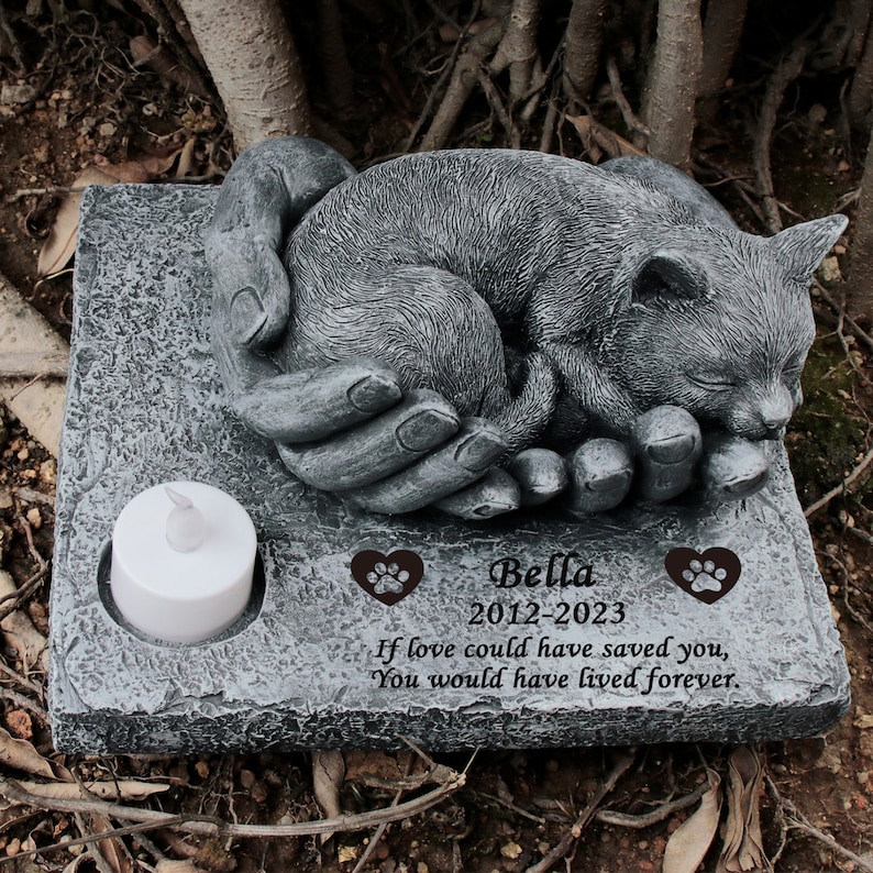 May include: A gray stone memorial with a sleeping cat sculpture in a pair of hands. The memorial has the inscription "Bella 2012-2023 If love could have saved you, You would have lived forever."