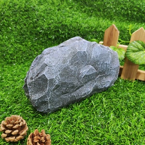 May include: A gray, textured rock with a rough surface. The rock is sitting on a bed of green artificial grass.