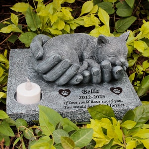 May include: A gray stone memorial with a sleeping kitten in hands. The memorial has a small candle holder and a personalized inscription that reads "Bella 2012-2023 If love could have saved you, you would have lived forever."