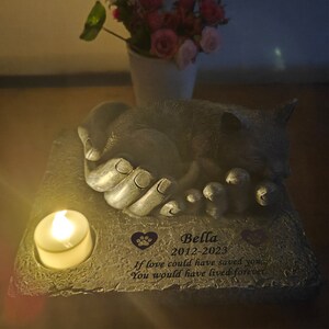 May include: A gray stone memorial with a sleeping cat sculpture. The memorial has a small candle holder and a personalized inscription that reads "Bella 2012-2023 If love could have saved you You would have lived forever".