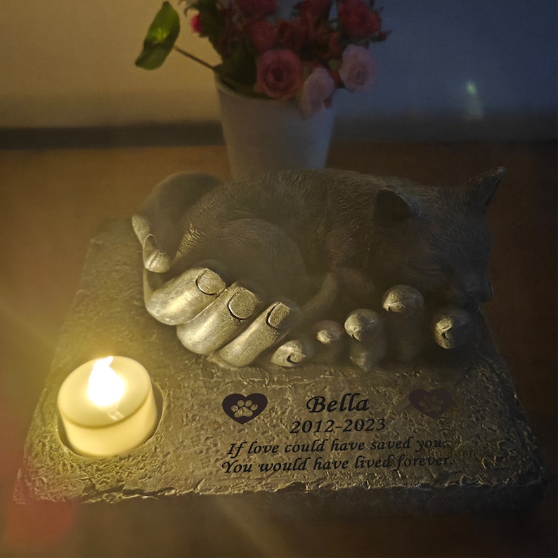 May include: A gray stone memorial with a sleeping cat sculpture. The memorial has a small candle holder and a personalized inscription that reads "Bella 2012-2023 If love could have saved you You would have lived forever".