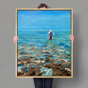 以下が含まれることがあります： 金色の額縁に入った長方形の油絵。白い帽子とシャツを着た人物が、浅いターコイズブルーの海の中に立っている様子が描かれています。前景は透明な水で、茶色と青の色調の岩の多い海底が透けて見えています。