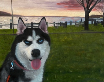 May include: A black and white husky dog with blue eyes sits on a grassy field with a red lead attached to its collar. The dog is looking at the camera with its tongue sticking out. The background shows a body of water with a sunset sky and a tree in the distance.