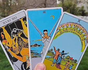 May include: Three tarot cards are fanned out, featuring vibrant illustrations. The cards include "THE DEVIL" with a yellow and black design, "II" with crossed swords, and "X" with a colorful arc. The background is a blurred outdoor scene.
