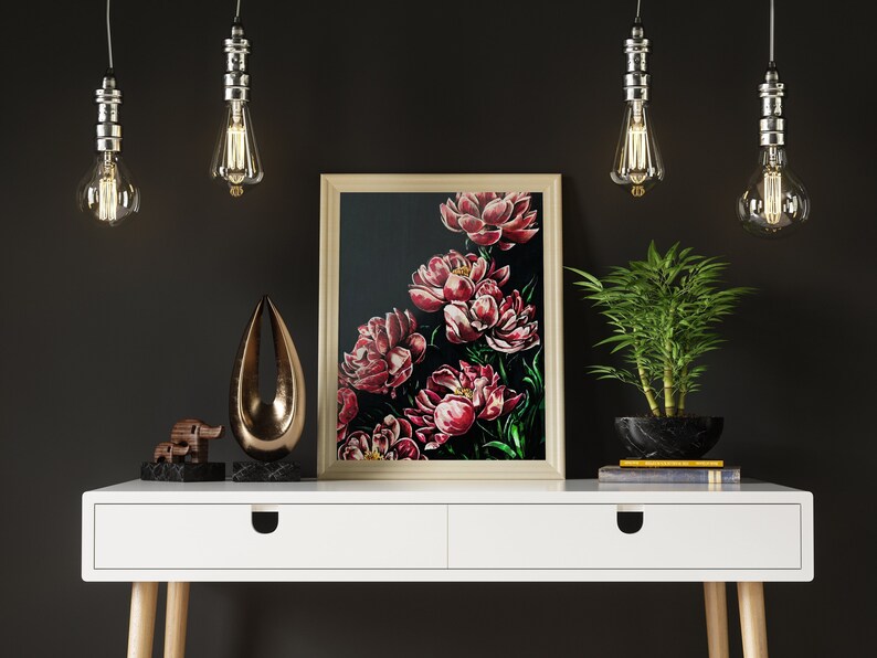May include: A white desk with two drawers and wooden legs, featuring a framed painting of red peonies on a black background, a gold metal sculpture, a wooden elephant figurine, a potted green plant, and three hanging light bulbs.