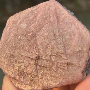 May include: A rough, pink gemstone with a textured surface and visible crystalline structures. The stone has a natural, unpolished appearance, showcasing its raw form. The color is a soft, dusty rose, with subtle variations in tone.