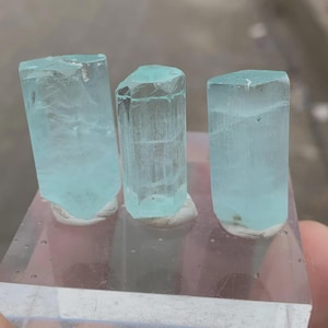 Puede incluir: Tres cristales de aguamarina azul claro se exhiben sobre una base de acrílico transparente. Los cristales tienen una apariencia translúcida con estructuras internas visibles. Los cristales son de diferentes tamaños y formas, mostrando formaciones naturales. El fondo está borroso.