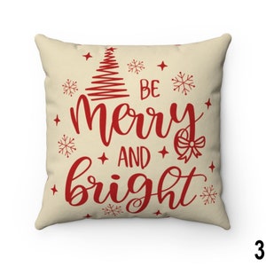 Pu&ograve; includere: Cuscino quadrato con sfondo color crema. Il cuscino presenta le parole "Be Merry and Bright" in corsivo rosso, insieme a un albero di Natale, fiocchi di neve e stelle. Un oggetto decorativo festivo per la casa.