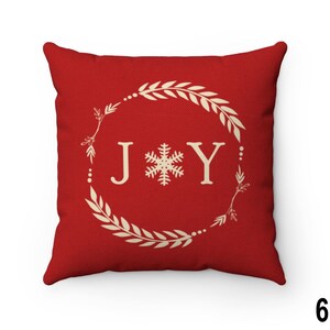 Pu&ograve; includere: Un cuscino decorativo rosso quadrato con la scritta "JOY" in bianco, con un fiocco di neve tra le lettere. Le lettere sono all'interno di una ghirlanda color crema con foglie e bacche. Un accento festivo per le festivit&agrave;.