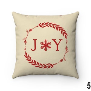 Pu&ograve; includere: Cuscino quadrato con sfondo color crema. Presenta un design a ghirlanda rossa con la parola "JOY" in lettere rosse e un fiocco di neve rosso al centro. Una decorazione festiva per le vacanze.