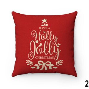 Pu&ograve; includere: Un cuscino quadrato rosso con la scritta "Have a Holly Jolly Christmas" in crema in un carattere decorativo. Il testo &egrave; disposto a forma di albero di Natale, con una stella in cima e un fiocco in basso.