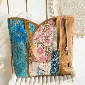 May include: A tote bag featuring a patchwork design of blue and pink floral fabric panels with white kantha stitching. The bag includes tan leather straps, a tan leather side panel with brass rivets, and a decorative silver-toned skeleton key charm.