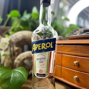 May include: A clear glass Aperol bottle with a blue and orange label. The label features the word "APEROL" in large, bold, yellow letters. The bottle is empty and sits on a wooden surface, with green foliage in the background.