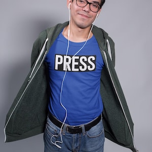Puede incluir: Una persona lleva una camiseta azul con la palabra "PRESS" en letras blancas sobre un rectángulo negro. Lleva una sudadera con capucha verde oscuro con cremallera, vaqueros azules y gafas. Hay auriculares blancos en sus oídos.