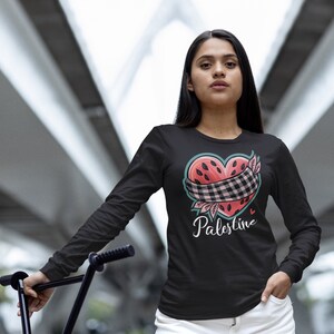 May include: A woman wearing a black long-sleeved shirt with a watermelon heart graphic and the word "Palestine" printed on it. She is wearing white denim shorts and holding a black BMX bike.