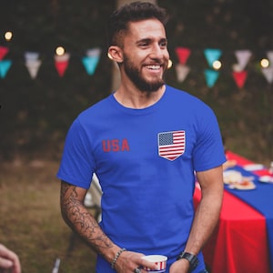 Peut inclure: Un t-shirt bleu avec le mot "USA" en rouge et un petit embl&egrave;me du drapeau am&eacute;ricain. La personne sourit. L'image est prise &agrave; l'ext&eacute;rieur.