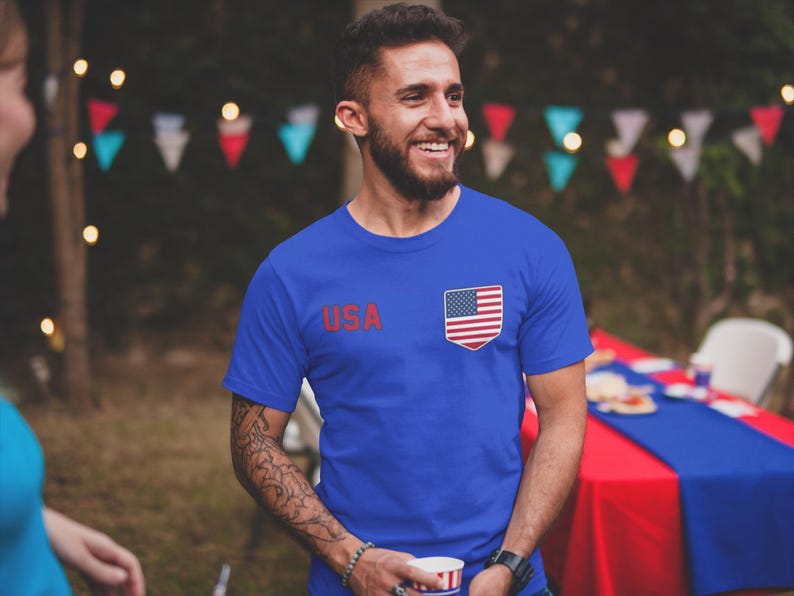 Peut inclure: Un t-shirt bleu avec le mot "USA" en rouge et un petit embl&egrave;me du drapeau am&eacute;ricain. La personne sourit. L'image est prise &agrave; l'ext&eacute;rieur.