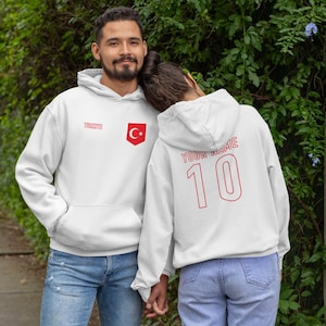 May include: Two white hoodies with red detailing. One displays the word "TURKIYE" and a shield with a crescent moon and star. The other has "YOUR NAME" and the number "10" on the back. Both are worn with blue jeans.
