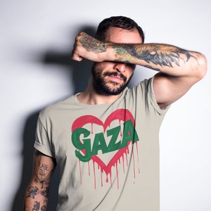 以下が含まれることがあります： 赤いハートのグラフィックと、ハートの中に緑色の文字で「GAZA」と書かれたベージュのTシャツ。ハートは赤い塗料が垂れている。