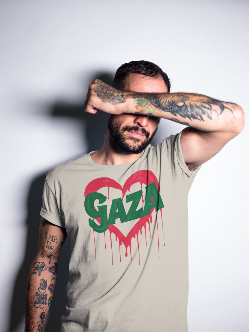 以下が含まれることがあります： 赤いハートのグラフィックと、ハートの中に緑色の文字で「GAZA」と書かれたベージュのTシャツ。ハートは赤い塗料が垂れている。