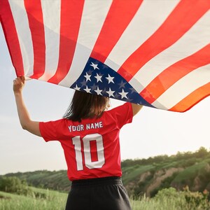 Peut inclure: Une personne tient un grand drapeau am&eacute;ricain &agrave; rayures rouges et blanches et un champ bleu d'&eacute;toiles blanches. La personne porte un maillot rouge avec le texte "YOUR NAME" et le num&eacute;ro "10" en blanc.