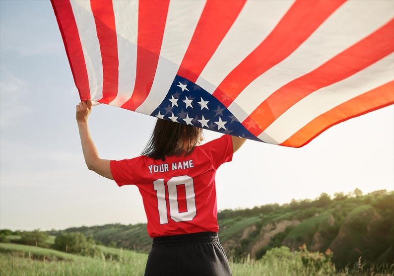 Peut inclure: Une personne tient un grand drapeau am&eacute;ricain &agrave; rayures rouges et blanches et un champ bleu d'&eacute;toiles blanches. La personne porte un maillot rouge avec le texte "YOUR NAME" et le num&eacute;ro "10" en blanc.