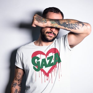 以下が含まれることがあります： 赤いハートのグラフィックと緑色の文字で「GAZA」と書かれた白いTシャツ。赤い塗料が垂れている。
