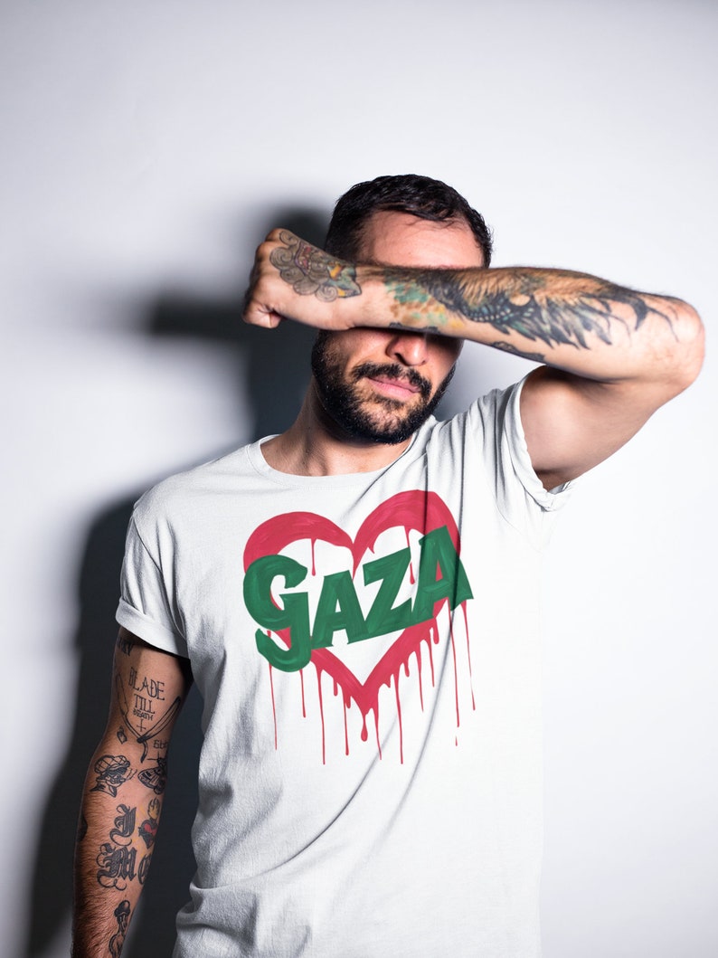 以下が含まれることがあります： 赤いハートのグラフィックと緑色の文字で「GAZA」と書かれた白いTシャツ。赤い塗料が垂れている。