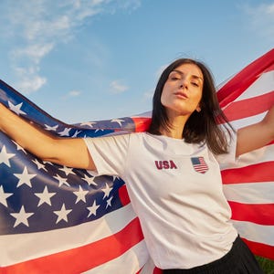 Peut inclure: Une personne tenant un grand drapeau am&eacute;ricain contre un ciel bleu. La personne porte un t-shirt blanc avec "USA" imprim&eacute; dessus, ainsi qu'un petit embl&egrave;me du drapeau am&eacute;ricain. Les rayures rouges et blanches et le champ bleu avec des &eacute;toiles blanches du drapeau sont bien en vue.