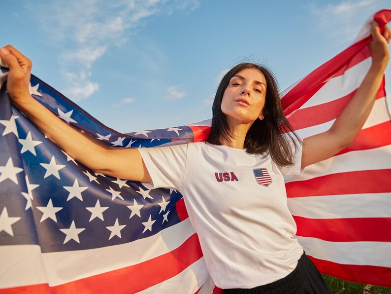 Peut inclure: Une personne tenant un grand drapeau am&eacute;ricain contre un ciel bleu. La personne porte un t-shirt blanc avec "USA" imprim&eacute; dessus, ainsi qu'un petit embl&egrave;me du drapeau am&eacute;ricain. Les rayures rouges et blanches et le champ bleu avec des &eacute;toiles blanches du drapeau sont bien en vue.