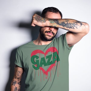 以下が含まれることがあります： 赤いハートの中に緑色の文字で「GAZA」と書かれた緑色のTシャツ。ハートは赤い塗料が垂れている。