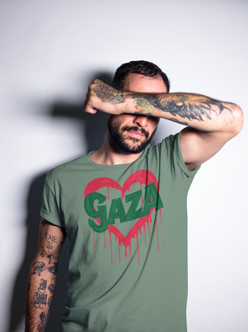 以下が含まれることがあります： 赤いハートの中に緑色の文字で「GAZA」と書かれた緑色のTシャツ。ハートは赤い塗料が垂れている。