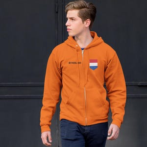 Könnte beinhalten: Ein orangefarbener Kapuzenpullover mit Reißverschluss, Kapuze und Vordertaschen. Der Hoodie zeigt den Text "NETHERLANDS" in Schwarz und ein niederländisches Flaggenemblem. Die Person trägt eine blaue Jeans.