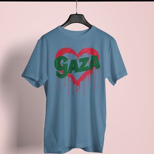以下が含まれることがあります： 薄い青色のTシャツに、緑色の塗料が垂れている赤いハートが描かれています。ハートの中に緑色の文字で「GAZA」と書かれています。