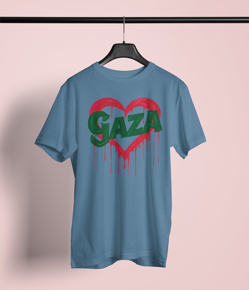 以下が含まれることがあります： 薄い青色のTシャツに、緑色の塗料が垂れている赤いハートが描かれています。ハートの中に緑色の文字で「GAZA」と書かれています。