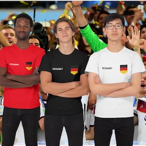 Op de afbeelding: Drie mensen dragen rode, zwarte en witte t-shirts met het woord "Germany" en een Duits vlagontwerp. Op de achtergrond juicht een menigte, met confetti.