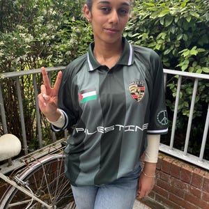May include: Dark green and grey striped football shirt with the word "PALESTINE" across the front. The shirt has a white collar and cuffs, and features the flag of Palestine and a team logo. Worn with blue jeans.