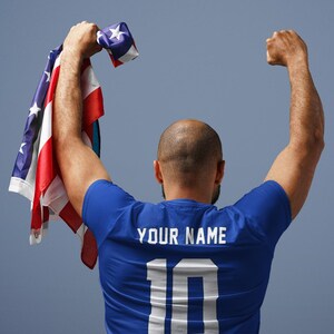 Peut inclure: Une personne portant un maillot bleu avec le texte "YOUR NAME" et le num&eacute;ro "10" dans le dos, tenant un drapeau am&eacute;ricain pli&eacute;. Les bras sont lev&eacute;s en signe de victoire sur fond bleu.