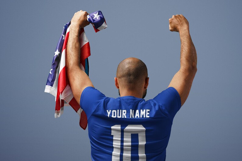 Peut inclure: Une personne portant un maillot bleu avec le texte "YOUR NAME" et le num&eacute;ro "10" dans le dos, tenant un drapeau am&eacute;ricain pli&eacute;. Les bras sont lev&eacute;s en signe de victoire sur fond bleu.
