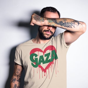 以下が含まれることがあります： 赤いハートの中に緑色の文字で「GAZA」と書かれたベージュのTシャツ。ハートは赤い塗料が垂れている。
