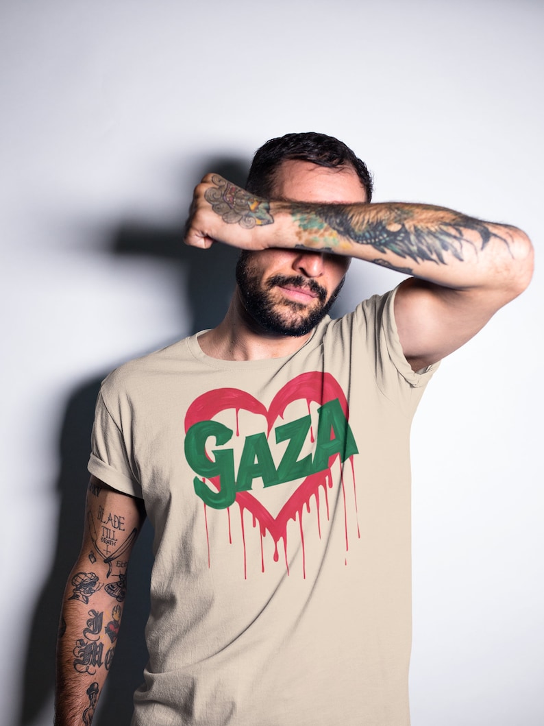 以下が含まれることがあります： 赤いハートの中に緑色の文字で「GAZA」と書かれたベージュのTシャツ。ハートは赤い塗料が垂れている。